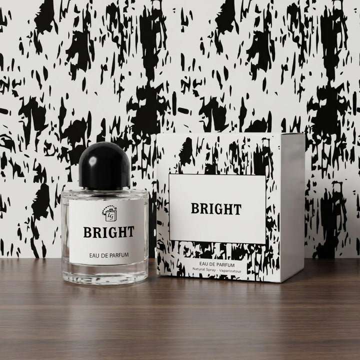 A clear glass perfume bottle with a black cap and a white label reading BRIGHT, placed beside its matching white and black patterned box on a wooden surface against a black-and-white abstract wall.